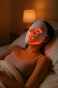Woman relaxing in bed wearing 7 color LED light therapy face mask with warm red glow - self-care skincare night routine